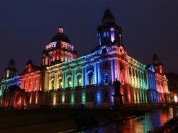Night time and the exterior of Belfast City Hill is lit up with pillars of gree, blue, purple, red and yellow light.